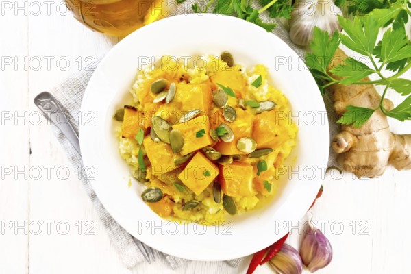 Millet porridge with spicy pumpkin sauce and seeds in a plate on a towel, hot pepper, ginger root, garlic and parsley on wooden board background from above