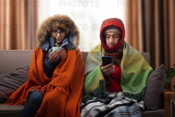 Middle age family in parkas drinking coffee on the sofa while using smartphone in their cold living room