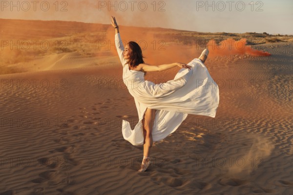 A girl in a fly white dress dances and poses in the sand desert at sunset