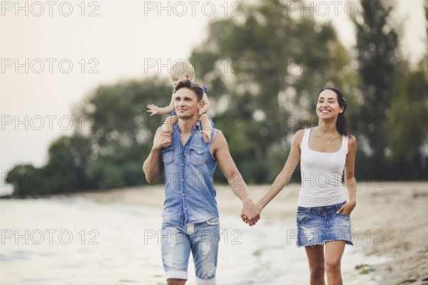 A young family of three walks along a sandy beach. A little baby girl with a blonde one year old from birth sits at the daddy on her shoulders, Dressed in blue jeans clothes
