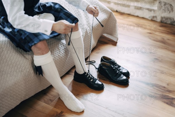 Preparing for a Scottish wedding. Man in a kilt, sporran and high socks sits on the bed and tie long shoelaces on his shoes.