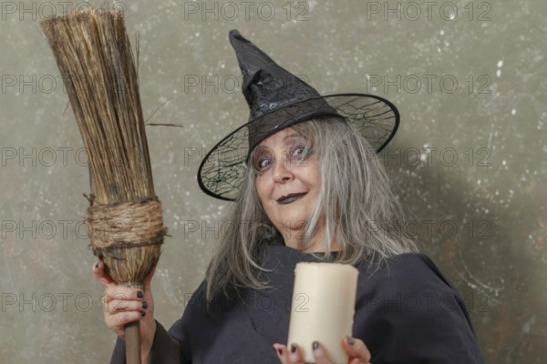 Old witch with white hair and a black hat holding a broom in her hand and a candle in the other while smiling