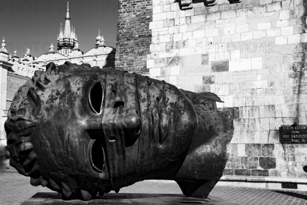 A large metal head sculpture lying on its side near a historical building in black and white, Sculpture, Igor Mitoraj, Krakow, Poland, Europe