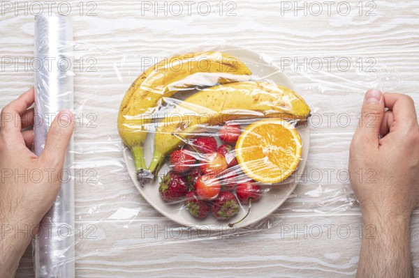 Male hands wrapping assorted different fruit and berries on white plate in plastic clear film top view on white wooden rustic table background, food photography, food photography