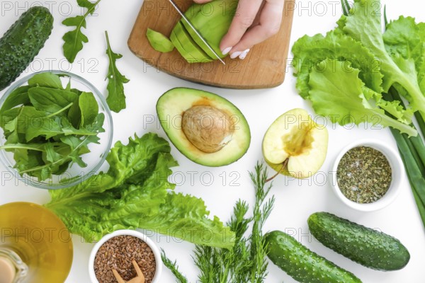 Woman cooking salad of fresh green vegetables and herbs. Cooking healthy diet or vegetarian food. Female hands cut avocado on cutting board surrounded by green vegetables. Step by step recipe. Healthy eating. Ingredients for salad. Pattern flat lay with healthy vegetarian meal ingredients. Raw food concept. A variety of organic fruits and vegetables with avocado. Vegan menu. Fresh spinach and green onion leaves