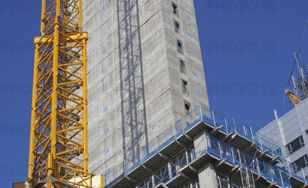 Close up of a large urban construction site with a yellow tower crane casting a shadow on a large concrete building and safety fences against a blue sky