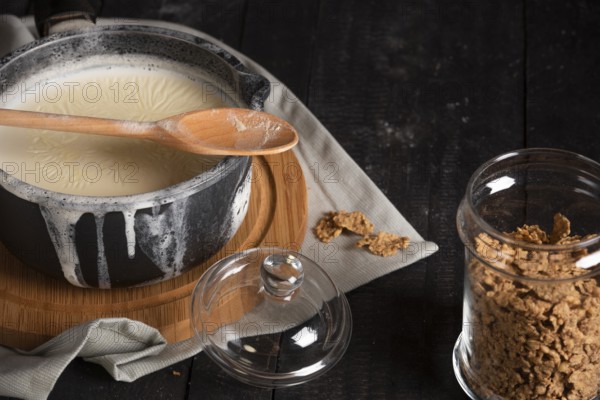 Whole grain cereals in a glass jar with lid and milk in a saucepan, spilled over the edges, on a rustic black table. Low light image. Healthy breakfast concept