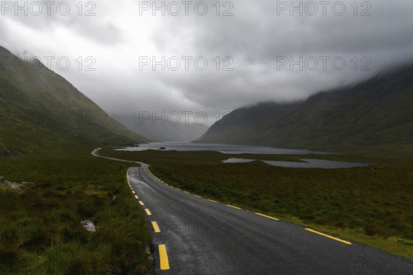 Narrow black-top highway with yellow road markings leads through an overcast mountain valley with fog and mist and lakes o nthe valley bottom