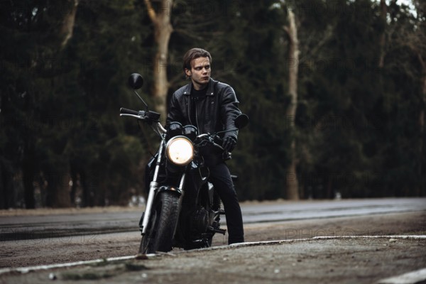 A person in a leather jacket sits on a motorcycle by a forest road, creating a moody atmosphere