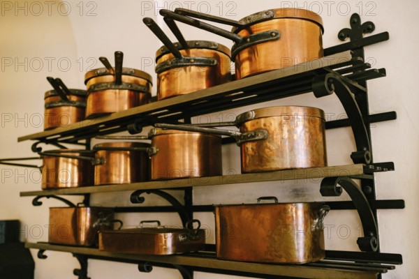 Pots, ladles, tray and saucepan made of copper, in various sizes, on massive metal shelves, fixed to a white wall.