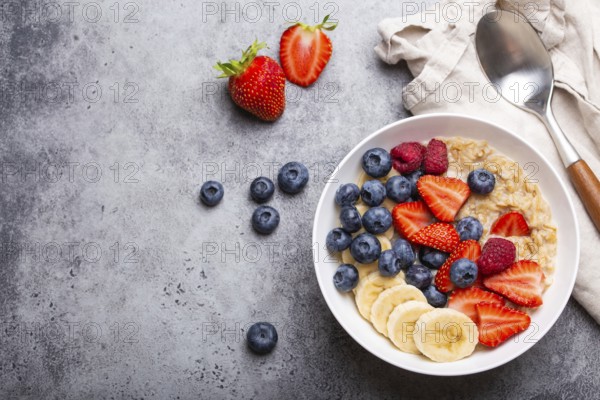 Oatmeal porridge with fruit and berries in bowl with spoon on gray stone background table top view, homemade healthy breakfast cereal with strawberry, banana, blueberry, raspberry. Space for text, food photography, food photography