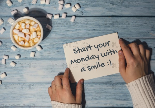 START YOUR MONDAY WITH A SMILE concept text Female hands holding card with white cup of coffee and marshmallows on wooden background. The most depressing day of the year in January. Day commit suicide and depression motivation sign. Top view, flat lay