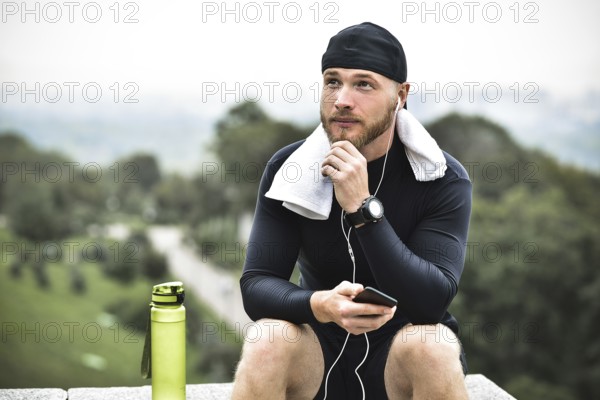 Muscular bearded athlete with towel checking burned calories on smartphone after good workout session on city park