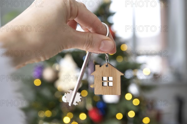 Key and tiny house keychain in hand on background fairy lights Christmas tree. gift for New Year, Christmas. Building, design, project, moving, insurance, mortgage, rent and purchase real estate