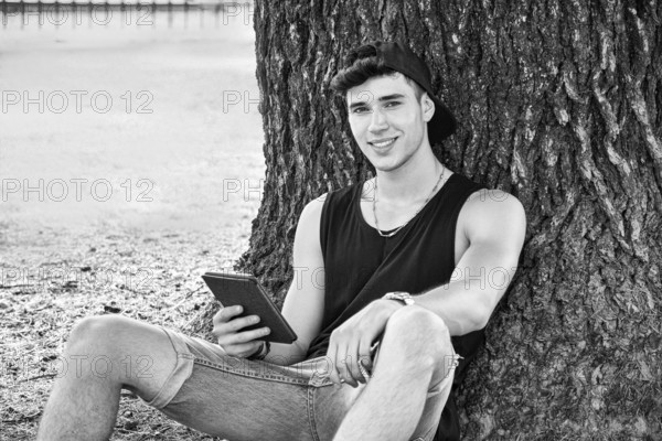 Smiling Young Handsome Man, Reading on Ebook Reader or Holding a Tablet Computer, Relaxing on Grassy Ground at Park, Leaning against Tree