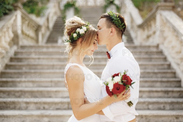 The bride and groom in wreaths stand hugging and kissing on the stairs of the ancient church in Prcanj, close-up.