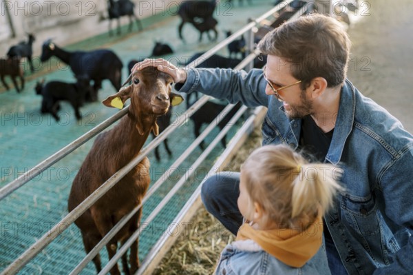Little girl stands next to her dad stroking a goat head poking out over a pen fence.