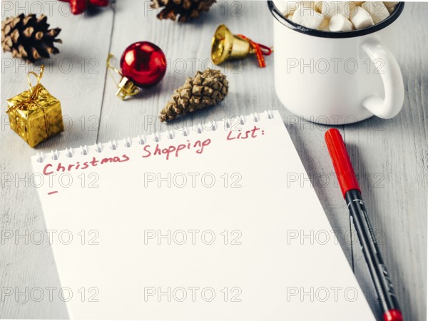 Christmas gifts shopping planning. Make shopping or to-do list for Christmas. Notebook, mug hot chocolate with marshmallows, New Year's decoration and pine cone on gray wooden background