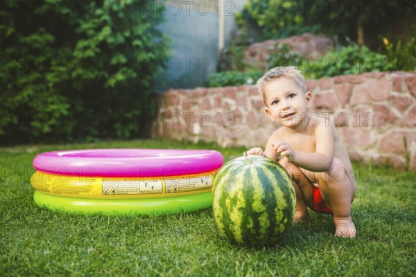 Toddler cute Caucasian white European race boy sitting hugging holding huge watermelon and smiling on background of green graß, backyard of the house and a round children pool on a sunny summer day