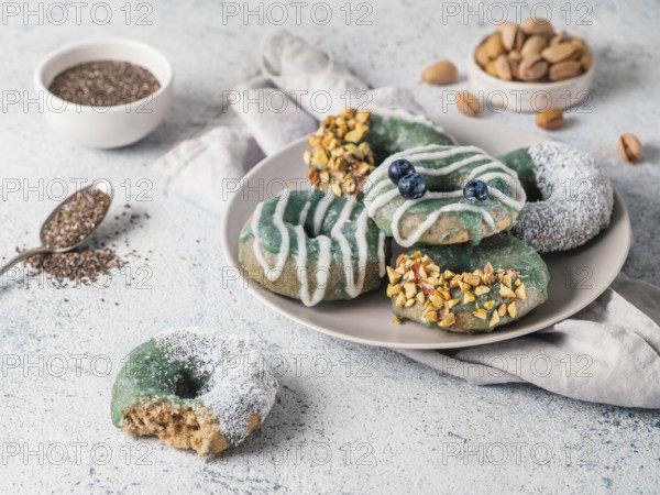 Vegan doughnuts with chia seeds topped with healthy spirulina glaze with pistachio, desiccated coconut and blueberry. Blue green spirulina donuts on gray background. Copy space