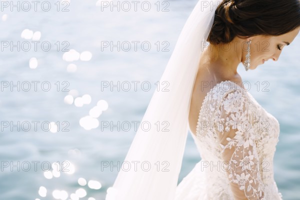 Close-up of the neck and neck of the bride, face profile. Beautiful big earring in ears, shoulder with white lace dress on background of glare of water. Fine-art wedding photo in Montenegro, Perast