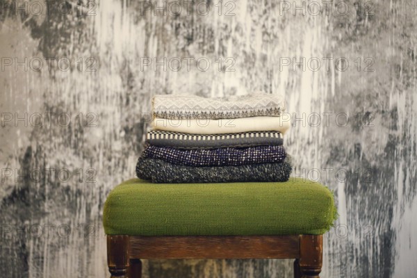 Pile of knitted warm grey and white blankets, scarves and woolen sweaters for winter or fall cold weather on white stool near white and gray wall, copy space. Cosy home winter or autumn atmospere