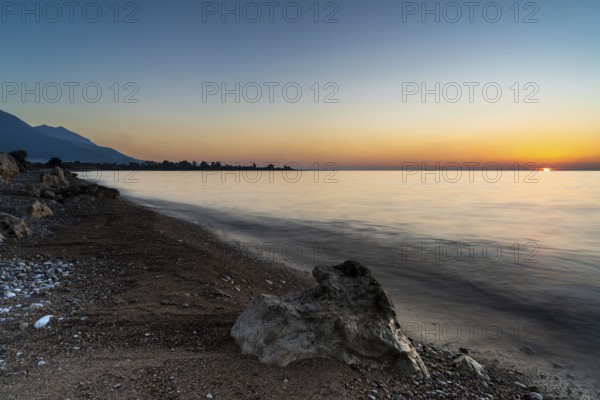 A peaceful sunset over the ocean with silhouette of mountains and pebble beach iwth rock boulders in the foreground