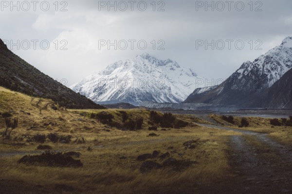 Gravel track leads through a vast landscape with snow-capped mountains and cloudy skies, Hooker Valley Track, New Zealand, Oceania