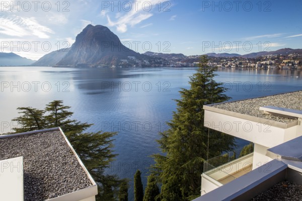 Fantastic park on coastline with giant spruce tree and cedar tree and apartment buildings (with white pebbles) on rooftop on coastlie. Mount San Salvatore on other side of lake Lugano and lugano, left part of Swiis city At left corner, part of dam