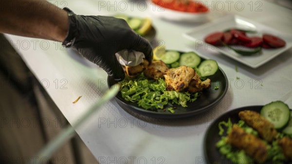 Gloved chef preparing a plate of chicken tikka garnished with fresh cucumber and lettuce in an Indian restaurant kitchen, ready to be served