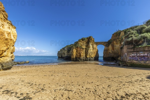 Yellow rocks and cliffs on a sandy beach. A bay by the sea, over which the old stone bridge of the Pinhão fortress connects two cliffs. Praia da Caldeira, Lagos. Algarve, Portugal, Europe