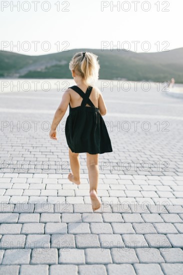 Little girl runs barefoot on a tiled road while hovering in the air. Back view.