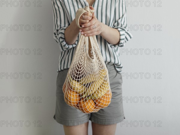 Mesh bag with fruits in female hands. Stylish young woman hold mesh shopping bag on light gray wall. Modern reusable shopping concept