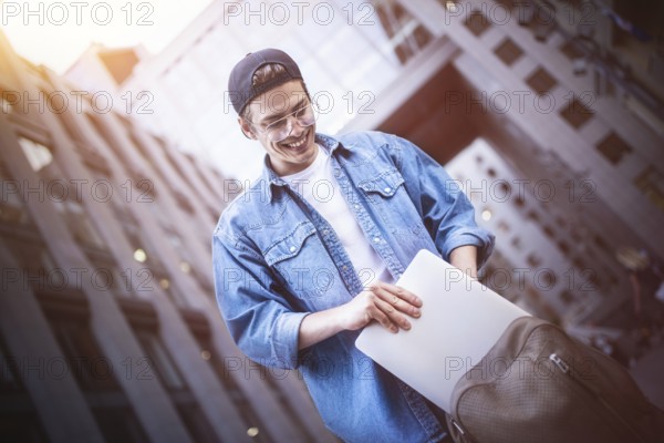 Happy handsome student looking at computer screen while typing something on laptop keyboard during work break outdoors, teenager looking at the notebook sitting at street