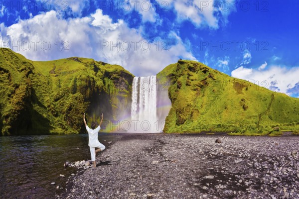 Elderly woman performs asana tree of hatha yoga before falling water. Grand waterfall with rainbow in the water mist Skogafoss