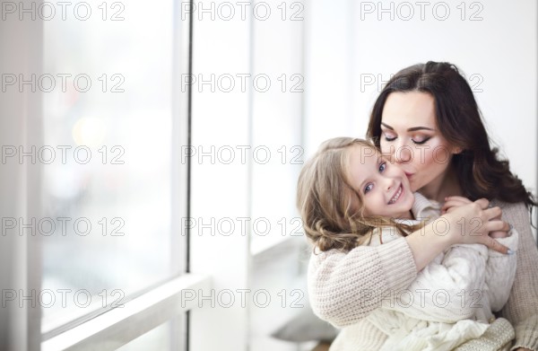 Happy loving family together. Mother and child daughter hugging by the window wearing cozy sweaters