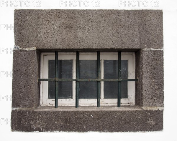 A small white wooden window with glass panes covered by steel bars in a large grey stone frame