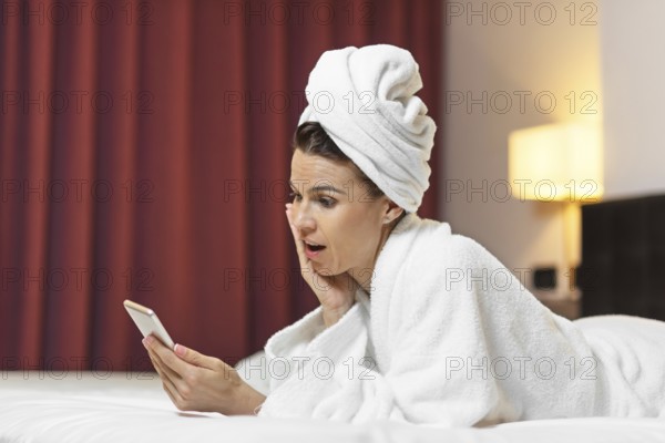 Young woman in bathrobe lying in hotel room using mobile phone, relaxed after taking a bath