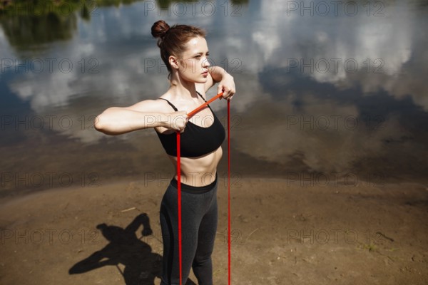 A focused woman exercises with a resistance band, casting a shadow on the riverbank
