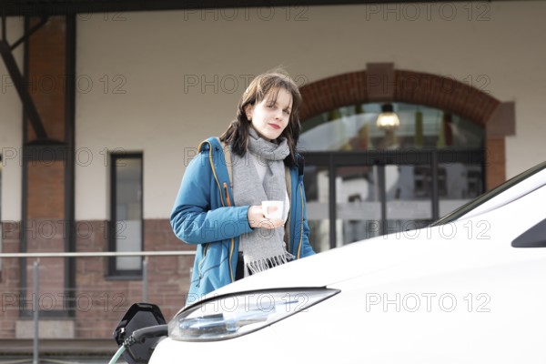 Woman with blue coat and cup in front of an electric car in urban environment in winter, eco-friendly