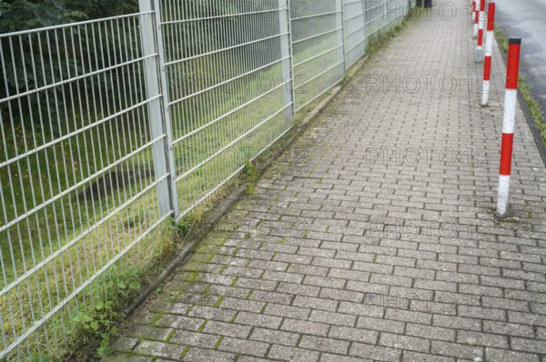 Sidewalk, metal fence on the left, red and white poles on the right