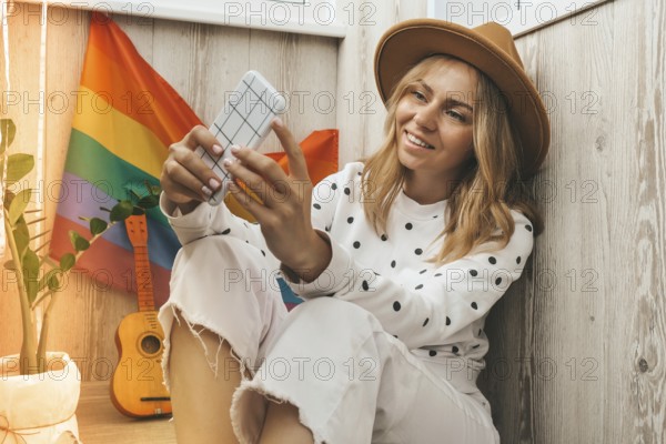 Young millennial hippie woman sitting on balcony using mobile phone. LGBTQ rainbow flag on background. Phone texting, video call, taking selfie. Blogger. Work from home. Modern office. Blonde hair. Stay home