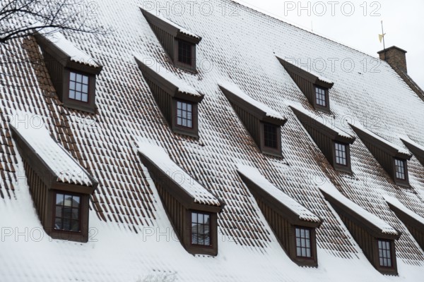 Snow falling on the roofs of the city. Snow-covered brown metal tiles roof of European house with windows. Rooftop covered in fresh snow. Winter scene