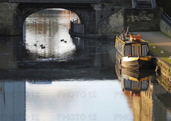 An old traditional narrow boat moored on the canal near brighouse basin in west yorkshire with ducks swimming under an old stone bridge
