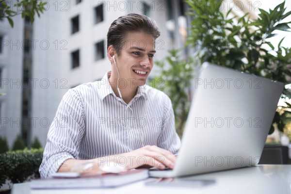 Happy handsome businessman looking at computer screen while typing something on laptop keyboard during work break outdoors, businessman with headphones looking at the notebook sitting in cafe working