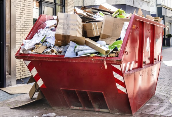 Red metallic container filled with cardboard trash in front of a shop downtown