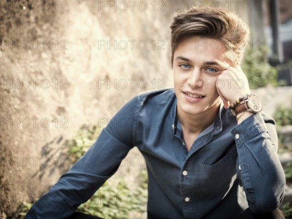 One handsome elegant young man in urban setting in European city, sitting in sunny day, looking at camera smiling happy