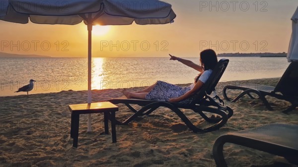 Young woman sitting on a sunbed at the beach pointing forefinger to the sunrise over the horizon. Beautiful female taking a sunbath at the sea. Summer vacation seaside, holiday relish concept