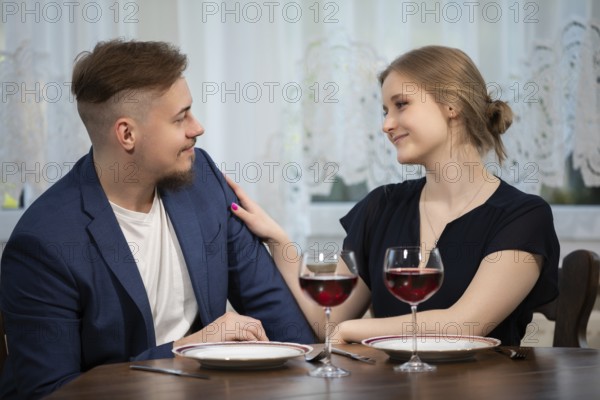 The girl strokes her partner's shoulder. The couple is sitting close together at a table. Two glasses of red wine stand on the table. The couple is elegantly dressed