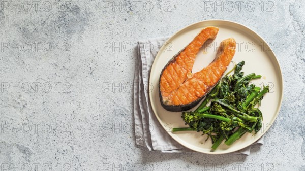 Ready-to-eat grilled salmon steak and greens - baby broccoli or broccolini and spinach on rustic craft plate over gray background. Keto diet dish. Top view or flat lay.Copy space left for text. Banner